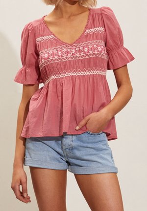 Vrouw die een roze blouse met pofmouwen en bloemenborduursel draagt en lichtblauwe opgerolde spijkershorts, staand met één hand in de zak.