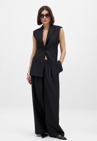 Chaleco negro a medida con solapa de muesca, diseño sin mangas y bolsillos laterales, combinado con pantalones de pierna ancha. Tejido mate con líneas marcadas.