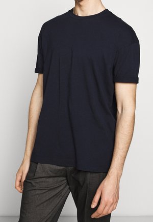 Granatowy bawełniany t-shirt z krótkimi wywiniętymi rękawami, okrągłym dekoltem i luźnym fasonem, zestawiony z szarymi spodniami.