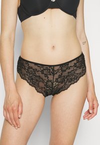 Svarte blonde truse med blomstermønster, med semi-transparent design og bølgete kanter. Myk tekstur og elastisk livband.