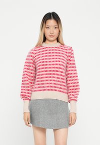 Sweter dzianinowy w jasno różowym kolorze z czerwonymi wzorami geometrycznymi, z prążkowanymi mankietami i dołem. W połączeniu z szaryą spódnicą.