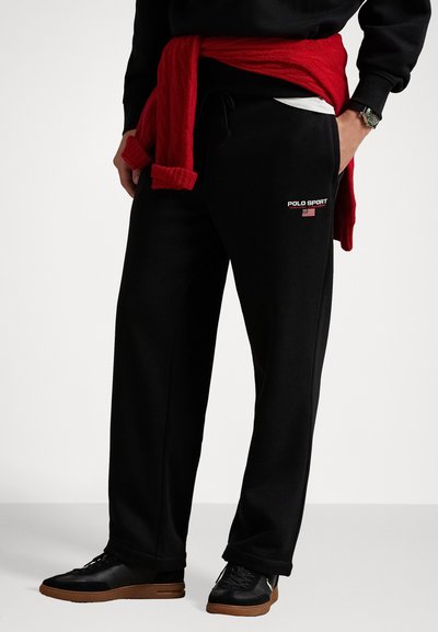 Eine Person trägt schwarze Polo Sport Trainingshosen mit Logo und amerikanischer Flagge, schwarze Sneakers, einen schwarzen Pullover und einen roten Pullover, der um die Taille gebunden ist.