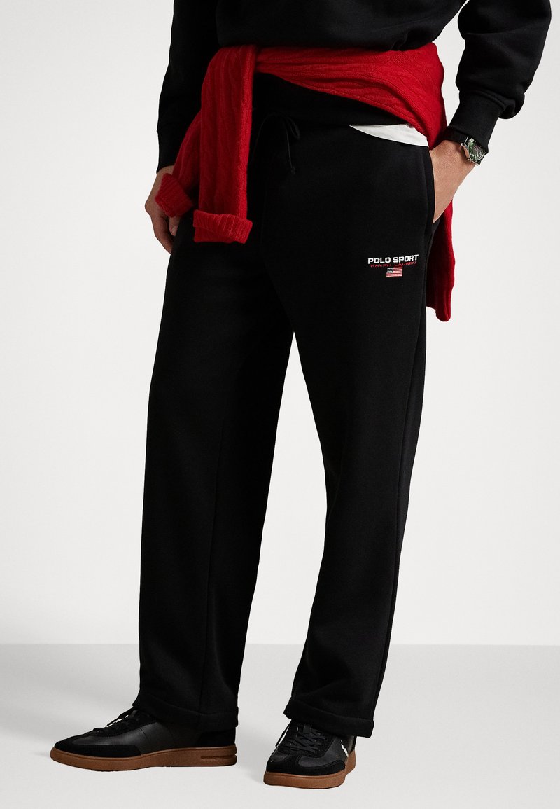 Persoană îmbrăcată în pantaloni de trening negri Polo Sport cu logo și steagul american, adidași negri, hanorac negru și un pulover roșu legat în talie.