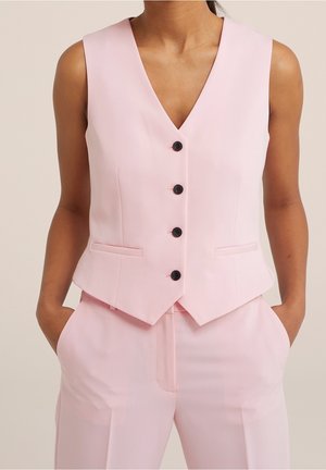 Femme portant un gilet sans manches rose clair avec des boutons noirs et un pantalon rose assorti, mains dans les poches, fond neutre.