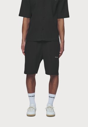 Personne portant une chemise noire à manches courtes avec boutons et un short, des chaussettes blanches hautes et des baskets blanches à lacets, debout devant un fond blanc.