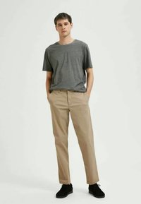 Hallhalli halli t-särk, kerge kanga ja beezide sirgete pükstega; mudel kannab musti jalanõusid, käed taskutes.