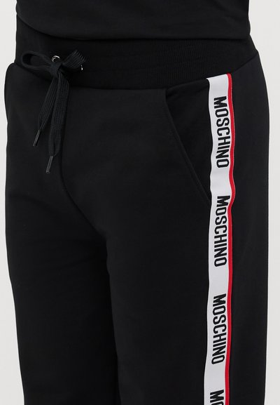 Crne trenirke s vezicom na struku, koje sadrže bočne pruge u crvenoj i bijeloj boji s ponovljenim natpisom "Moschino".