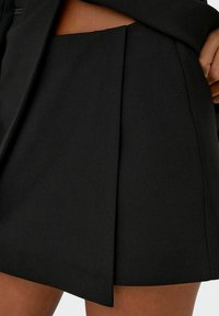 Zwarte geplooide rok van een gladde, gestructureerde stof. Heeft een hoge taille en een scherpe, asymmetrische zoom. Minimalistisch ontwerp en elegante snit.