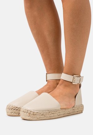 Beige espadrillot, joissa on nilkkahihna, sileä nahkainen päällinen, köysikudospohja ja tasainen muotoilu, korostaen minimalistista ja moderneja siluettia.