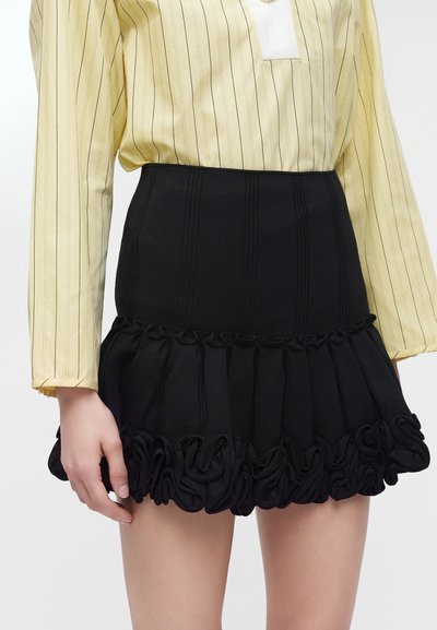 Femme portant une mini-jupe noire à volants assortie à un chemisier jaune à manches longues avec de fines rayures verticales.