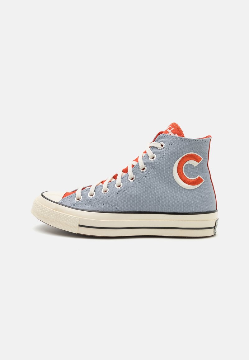 Basket montante en toile bleu clair avec lacets blancs, doublure intérieure rouge, logo rouge "C" sur le côté, et semelle en caoutchouc crème à rayures noires.