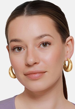 Donna con capelli castani tirati indietro, che indossa grandi orecchini a cerchio dorati e lucidi e una maglietta lilla chiaro, rivolta in avanti con un'espressione neutra.