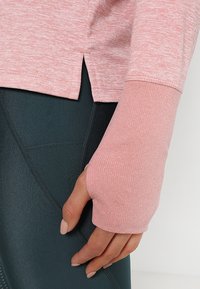 Langärmliges, hellrosa Sportoberteil mit gerippten Bündchen, seitlichen Schlitzen und einer weichen, strukturierten Oberfläche, kombiniert mit dunkelgrünen Leggings.