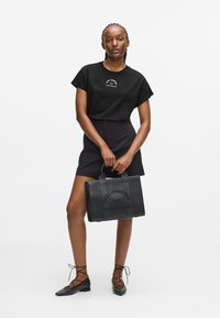 Fekete póló logóval, magas derekú fekete rövidnadrág, és egy texturált fekete kézitáska dombornyomott felirattal. Lapos fekete, tüskés cipők.