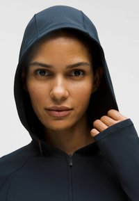 Sötétkék, cipzáros kapucnis pulcsi, illeszkedő kapucnival, sima textúrával és hüvelykujjlyukakkal. Minimalista varrás és letisztult dizájn jellemzi.