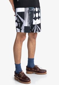 Shorts de bain noirs et blancs à motifs avec des designs géométriques et des formes contrastées, assortis à des chaussures bateau en cuir marron et des chaussettes bleues.