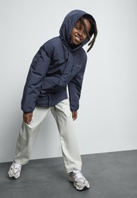 Smiling child with dreadlocks wearing a navy hooded jacket, light beige pants, and white sneakers, leaning forward on a gray floor.