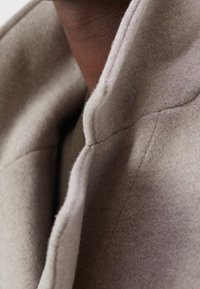 Manteau en mélange de laine doux gris clair avec un col haut et des coutures soignées, illustrant un design minimaliste avec une forme structurée.
