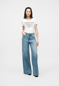 Νεαρή γυναίκα με ίσια μαύρα μαλλιά φοράει λευκό t-shirt με το λογότυπο της Guess και ψηλόμεσα, φαρδιά μπλε τζιν παντελόνια, στέκεται με το χέρι στην τσέπη.