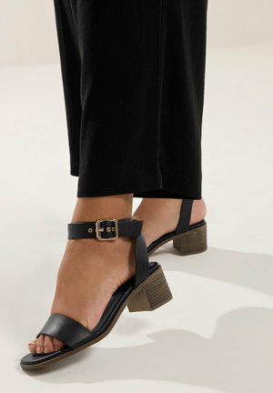 Voet met zwarte sandaal met lage blokhak, gouden gespsluiting en open teen, gecombineerd met zwarte broek tegen een lichte achtergrond.