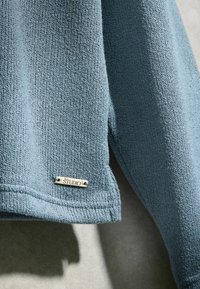 Manche de pull en maille bleu clair avec une petite étiquette métallique rectangulaire portant la mention "STUDIO" cousue près de l'ourlet sur un fond gris.