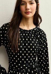 Vestido negro con escote redondo, detalles con pliegues y mangas largas. Presenta lunares blancos en forma de diamante y un cinturón a juego.