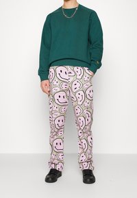 Sudadera verde de algodón, combinada con pantalones rosas que presentan un divertido patrón de caritas sonrientes. Unos zapatos negros completan el atuendo.