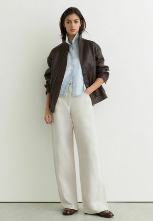 Vrouw in een bruine leren jas, lichtblauw overhemd met knopen, wijde witte broek en bruine schoenen staat tegen een witte muur.