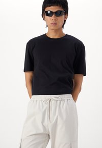 Schwarzes Baumwoll-T-Shirt mit kurzen Ärmeln, kombiniert mit hellbeigen Hosen mit elastischem Bund. Das Model trägt schwarze Sonnenbrillen und steht mit verschränkten Armen.