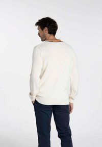 Pull en maille blanche à manches longues, avec des poignets côtelés et un col de couleur contrastante. Porté avec un pantalon sombre, vu de derrière.