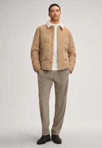 Giacca trapuntata beige con colletto in pelliccia sintetica bianca, abbinata a pantaloni grigi chiari e stivali alla caviglia marrone scuro. Presenta chiusura con bottoni e tasche.