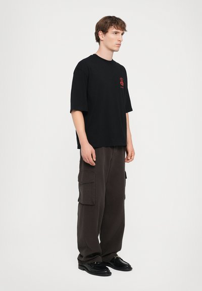 Schwarzes Baumwoll-T-Shirt mit lockerer Passform, kurzen Ärmeln und rotem grafischem Detail auf der Brust. Kombiniert mit lockeren braunen Cargo-Hosen.