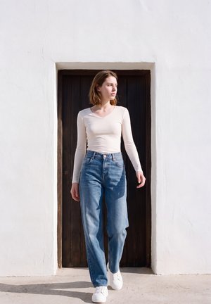 Une femme prend la pose, les mains dans les poches. Elle porte un haut blanc, un jean bleu à jambes larges et des baskets blanches, dans une embrasure de porte sombre.