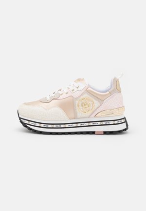 Zapatilla de deporte en rosa claro y beige con cordones blancos, logo dorado en el lateral, suela texturizada y franja de marca a lo largo de la suela exterior negra con dibujo.