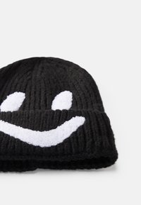 Zwarte gebreide beanie met een ontwerp van een witte glimlachende gezicht, textuurrijke geribbelde stof en een omgeslagen rand. Zacht en gezellig materiaal.