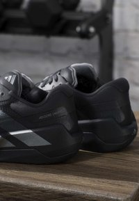 Ζευγάρι μαύρα παπούτσια τρεξίματος Adidas Adizero Dropset σε ξύλινη επιφάνεια με θολό εξοπλισμό γυμναστηρίου στο φόντο.