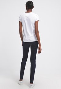 Camiseta blanca de manga corta con cuello redondo, combinada con jeans ajustados oscuros y zapatillas plateadas, vista desde atrás.