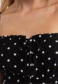 Närbild av en kvinna som bär en svart klänning med vita prickar och en knuten sløyfa vid den insamlade halsringningen.