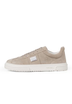Beige suède lage sneaker met witte rubberen zool, veters aan de voorkant en een rechthoekige merklogo patch aan de zijkant.
