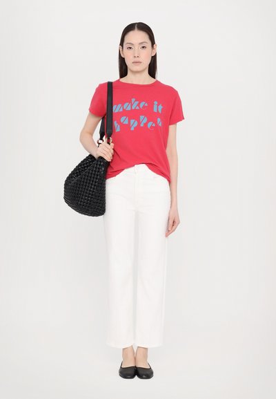 Red t-shirt with blue text design, white wide-leg jeans, black woven handbag, and black flats, standing against a white background.