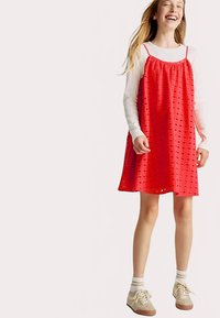 Rode oogjesjurk met een losse pasvorm, verstelbare bandjes en subtiele vierkante patronen; gedragen over een wit T-shirt met lange mouwen en gecombineerd met beige sneakers.