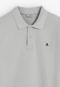 Polo de color gris claro con cuello clásico, tres botones y un pequeño logo de calavera y tibias en azul marino en el lado izquierdo del pecho. Textura suave.