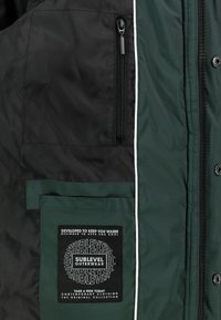Schwarz-grüne Jacke mit einer Reißverschlusstasche, weißer Paspelierung und einem großen schwarzen Label mit weißem Text auf der linken Brustseite.