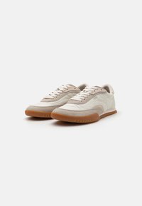 Beige treningssko med en strukturert overdel som kombinerer semsket skinn og syntetisk materiale. Gummi yttersåle med mønstrede spor og snøring.