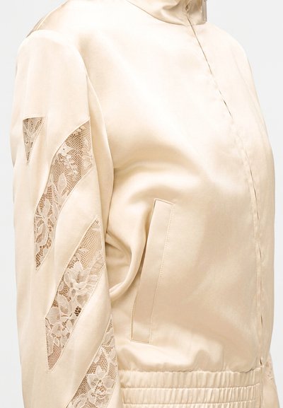 Veste en satin beige avec insertions en dentelle sur la manche, fermeture éclair devant, poche latérale et taille élastiquée, vue de côté.