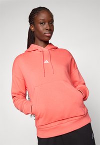 Korallfärgad hoodie med en framficka, justerbar dragsko och vit logotyp. Mjuk, texturerad tyg med avslappnad passform.
