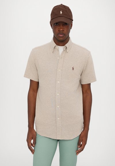 Camisa de botones de manga corta beige con un pequeño logo marrón en el pecho, combinada con una gorra marrón y pantalones verde claro.