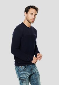 Marineblauer Strickpullover mit strukturiertem Körper, gerippten Bündchen und rundem Halsausschnitt. Kombiniert mit ausgewaschenen Jeans mit mehreren Taschen.