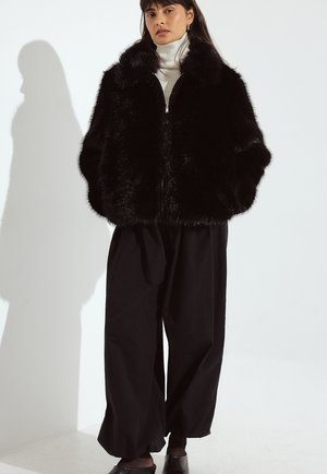 Sort faux fur-jakke med korte ærmer og lynlås, båret over en hvid turtleneck, parret med løse sorte bukser og sko.