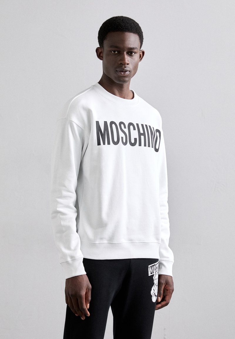 Sudadera blanca con el atrevido logotipo negro "MOSCHINO" en el pecho. Cuello redondo, puños y dobladillo acanalados. Llevada con pantalones negros que presentan un gráfico.
